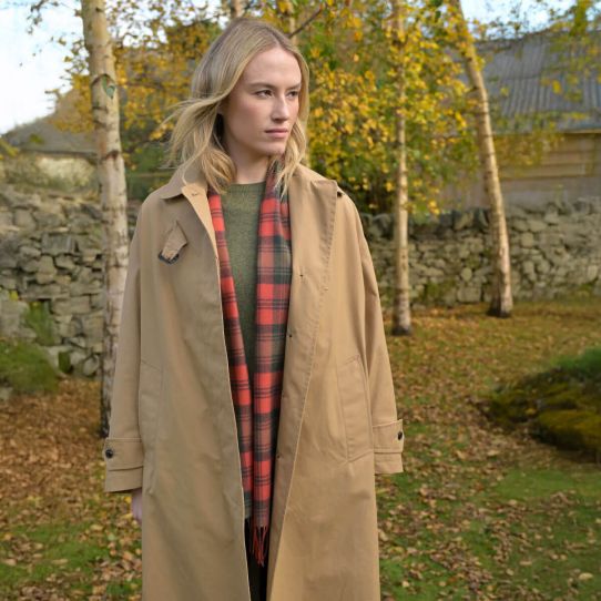A female model in the forest wearing a trench coat and green knitted jumper with a tartan scarf loosely hanging around her neck.