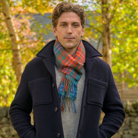 A man wearing a dark navy wool coat over a grey knit sweater, styled with a red, teal, and green plaid scarf, standing outdoors in front of a blurred background of autumn trees.