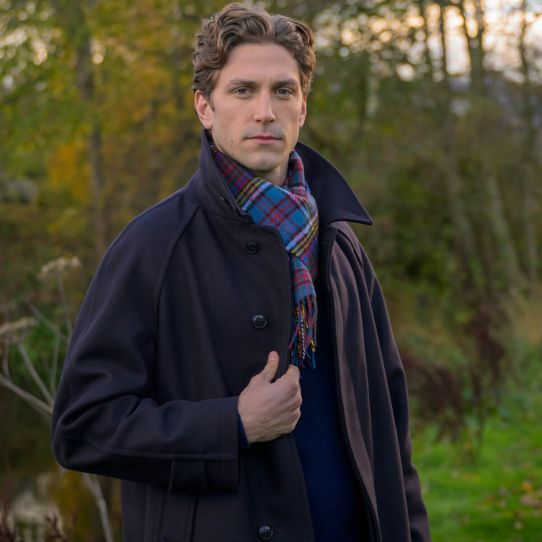 A close up of a man in the forest with a black coat with a lambswool colourful tartan print scarf in blue, red and yellow.