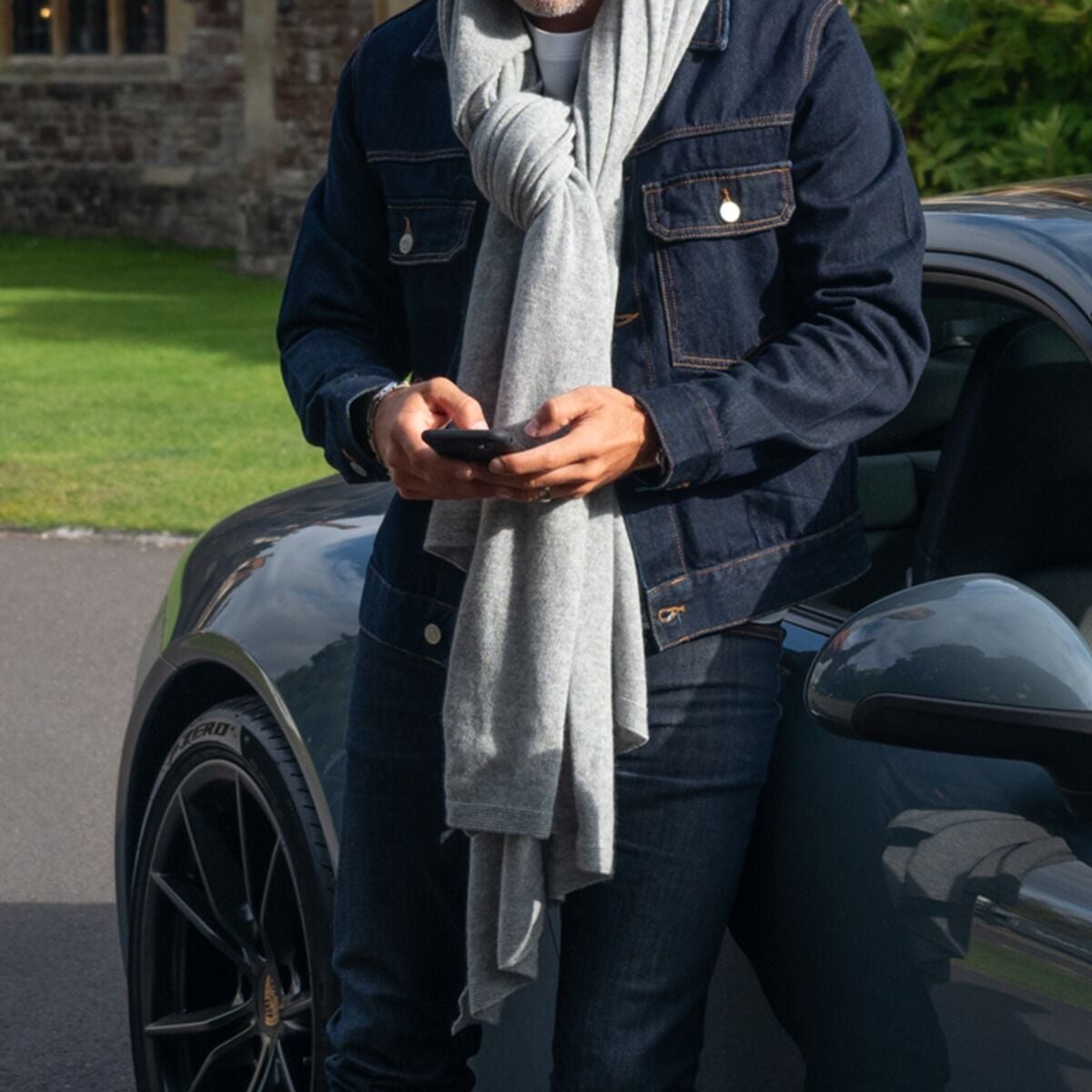 A person standing beside a dark car outdoors, wearing a dark denim jacket, dark jeans, and a light silver grey knit cashmere scarf while holding a smartphone.